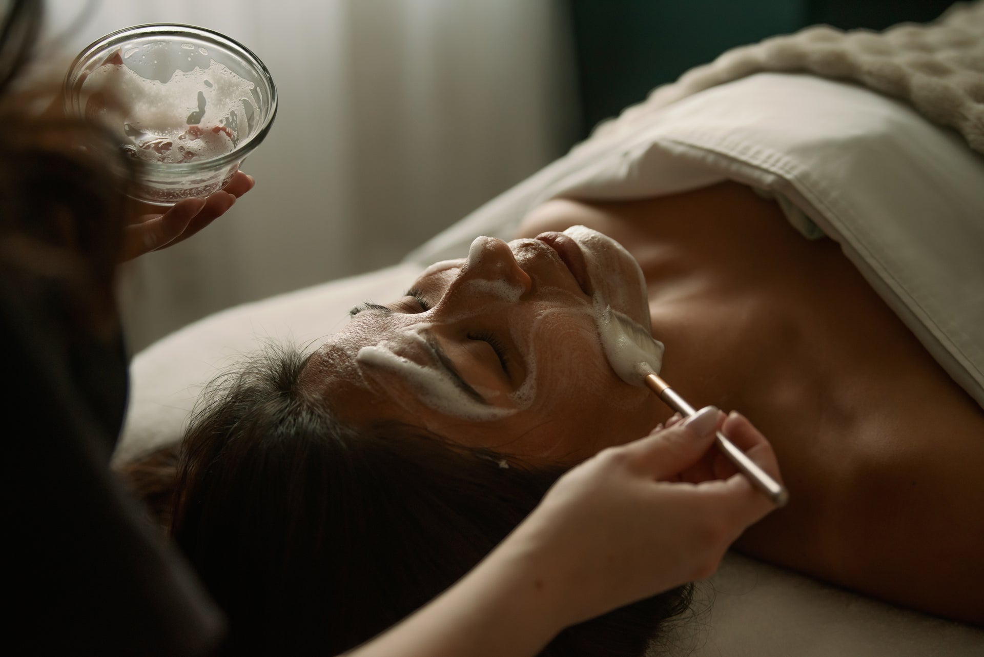 Esthetician brushing a custom mask onto a client's face from a glass mixing bowl