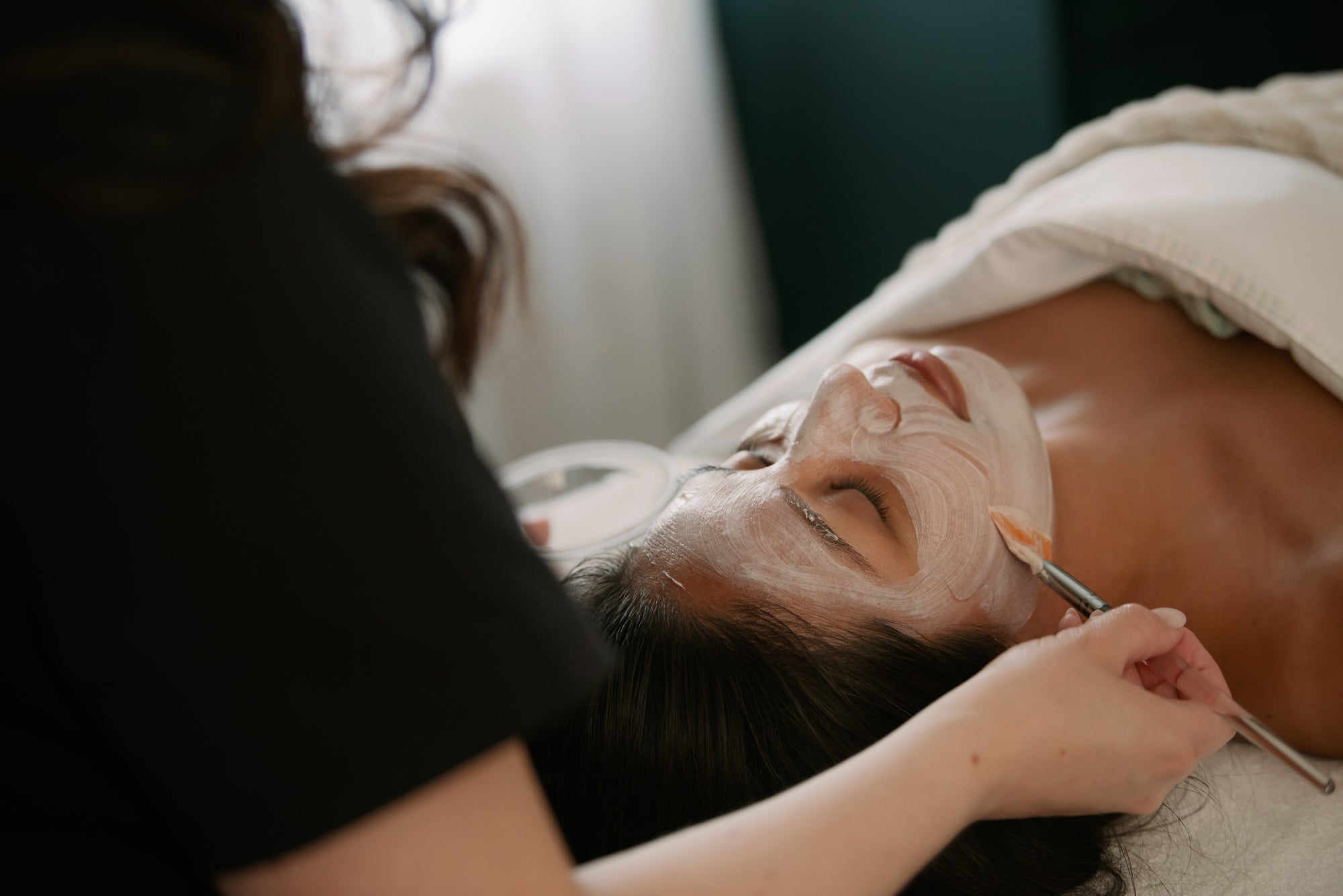 Esthetician brushing a treatment mask across a client's face in the dimly lit treatment room