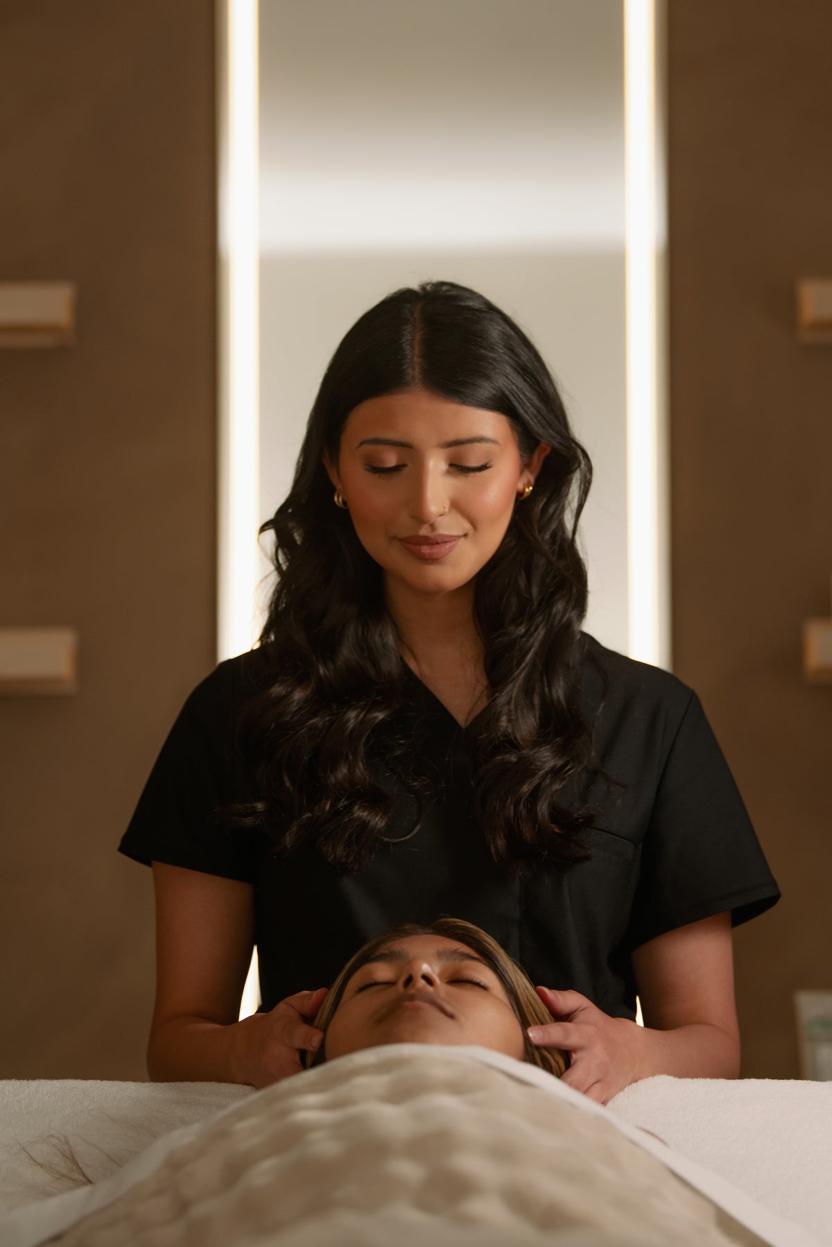 Esthetician beginning a facial at the head of the treatment bed beside a lighted mirror