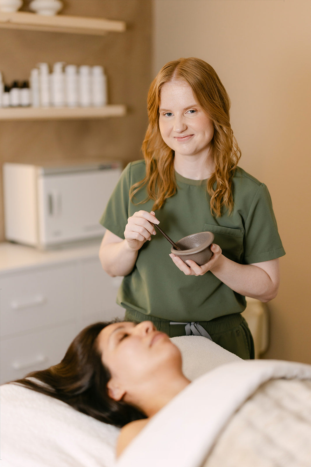 Esthetician holding a freshly mixed treatment mask beside a client resting on the facial bed