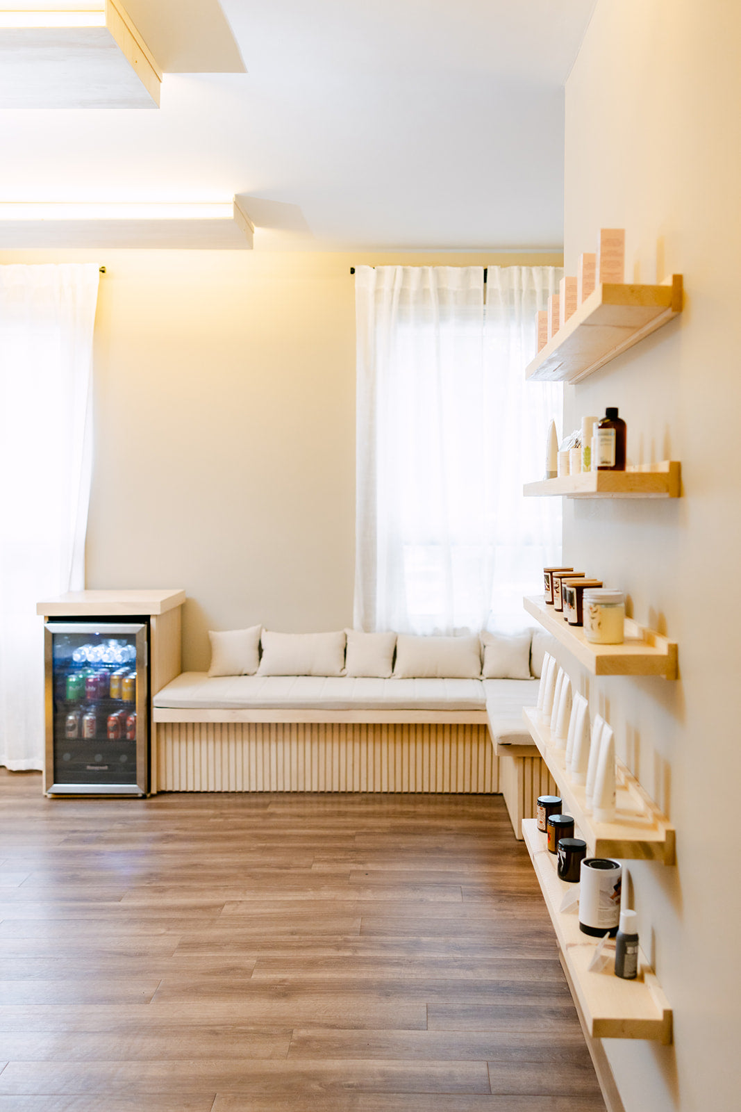 Spa sitting area with a bench sofa, sheer curtains, and retail shelves at Esthetics With Me